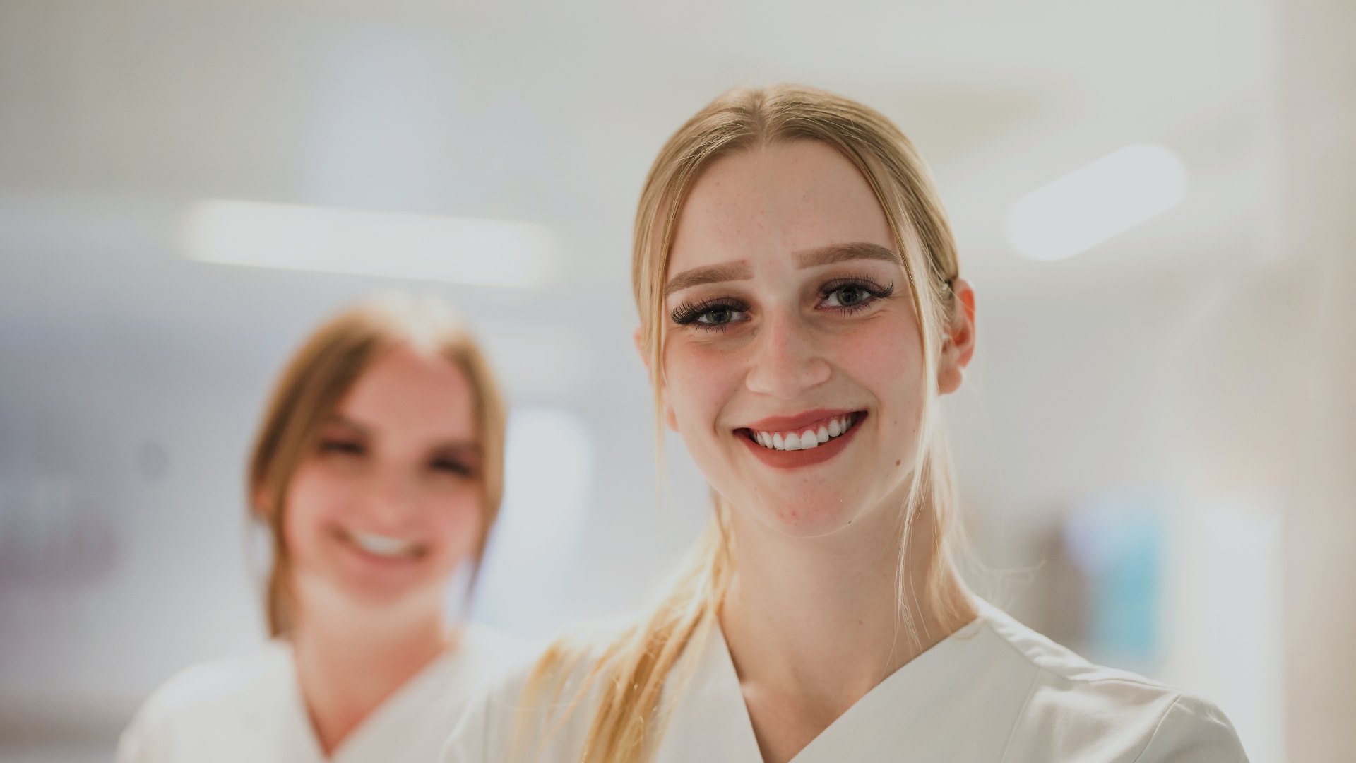 Junge Pflegerin rechts gro&szlig; im Bild mit blondem Pferdeschwanz l&auml;chelt in die Kamera; links dahinter Kollegin, leicht verschwommen